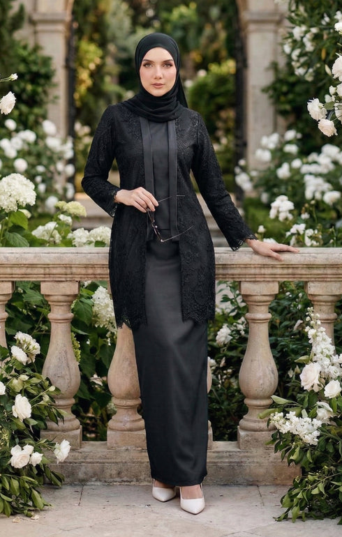 Woman in modern kebaya Kutu Baru Black with Instant Shawl hijab and outfit standing in a garden with white flowers and stone balustrade.