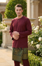 Baju Melayu Teluk Belanga Maroon front view full traditional outfit standing outdoors with greenery and flowers in the background.