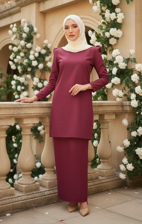 Woman in a Kurung moden plain maroon full outfit view dress standing in front of floral decorations.