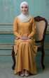 Woman in a Kebarung Klasik Plain Mustard and white plain hijab sitting on a wooden chair against a teal wall.