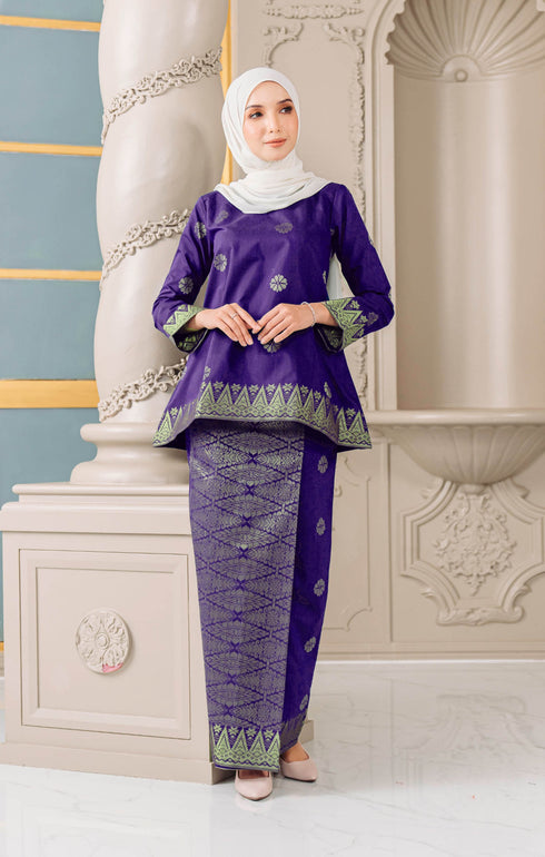 A woman wearing a traditional Kurung Kedah Purple Gold set with a round neckline and long sleeves, standing in a beautifully decorated room.