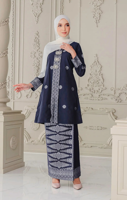 Woman wearing a Kebaya Blue Black Silver showing premium songket floral pattern traditional outfit with pearl white shawl in an ornate room.