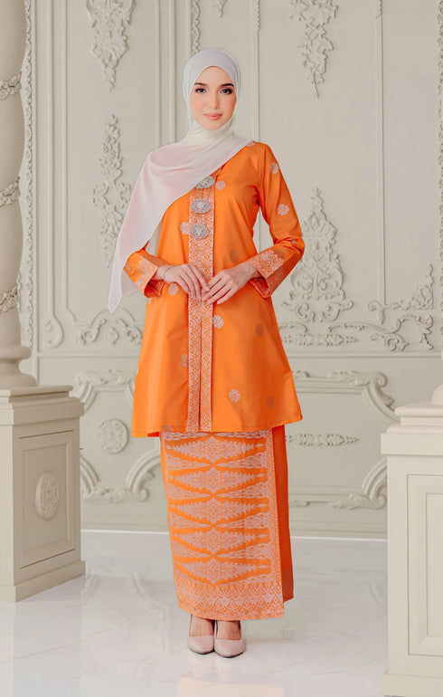 Woman wearing a Kebaya Orange Silver showing songket floral pattern and fit traditional outfit with a pearl white shawl hijab in a decorative room.