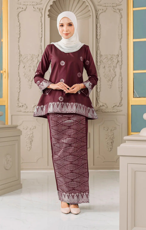 Woman wearing a traditional Kurung Kedah Dark Maroon Silver and white outfit with a matching shawl hijab in an elegant interior setting.