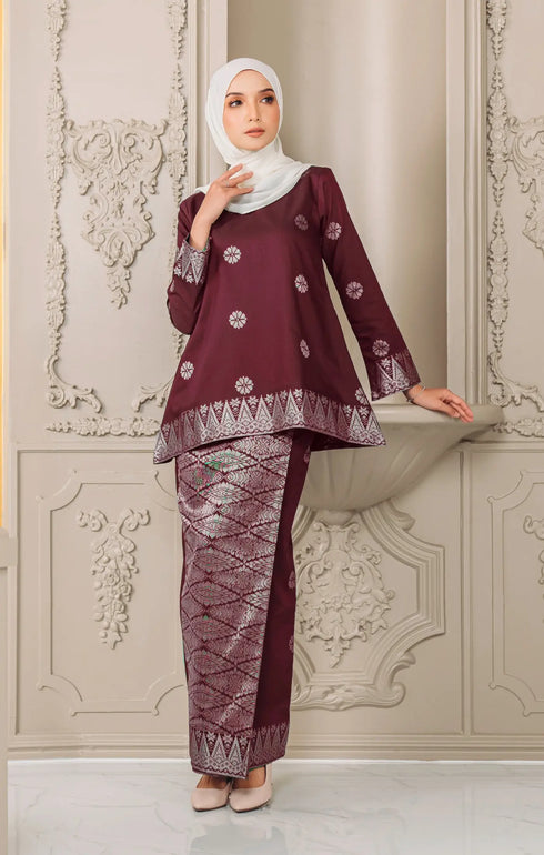 Woman wearing a traditional Kurung Kedah Dark Maroon Silver and white outfit with a instant tudung hijab against an ornate wall.