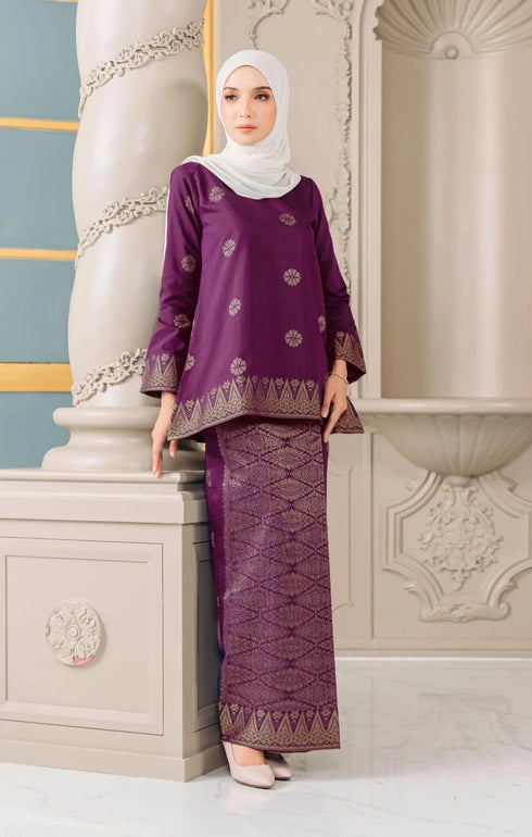 Woman wearing a Kurung Kedah Magenta Gold traditional outfit standing against an ornate architectural background.