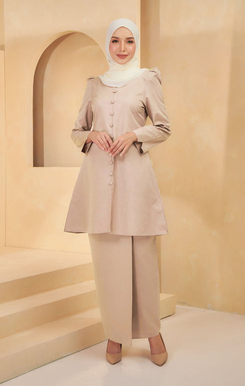 A woman wearing a Kurung Riau Plain nude colored with a V-neck top and long straight sleeves, paired with matching trousers. She is standing in a room with a neutral background.